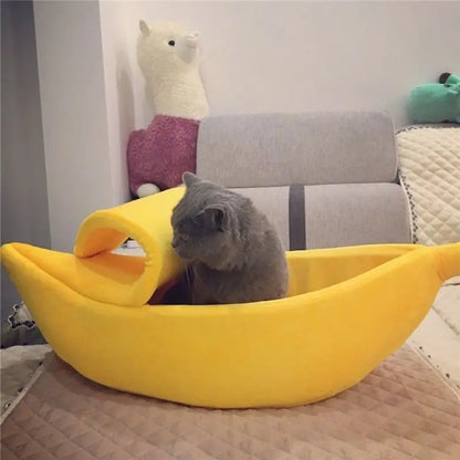 Cat lying inside a yellow banana-shaped pet bed with plush toys in the background.