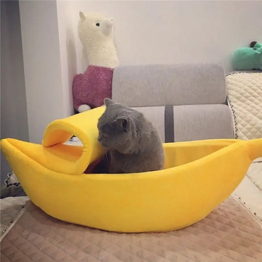 Cat lying inside a yellow banana-shaped pet bed with plush toys in the background.
